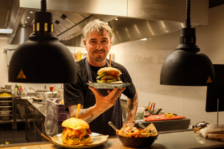 De chef van Wereldburgers aan het Akerkhof in Groningen presenteert een huisgemaakte burger in de open keuken, gefotografeerd voor Oogst Groningen.