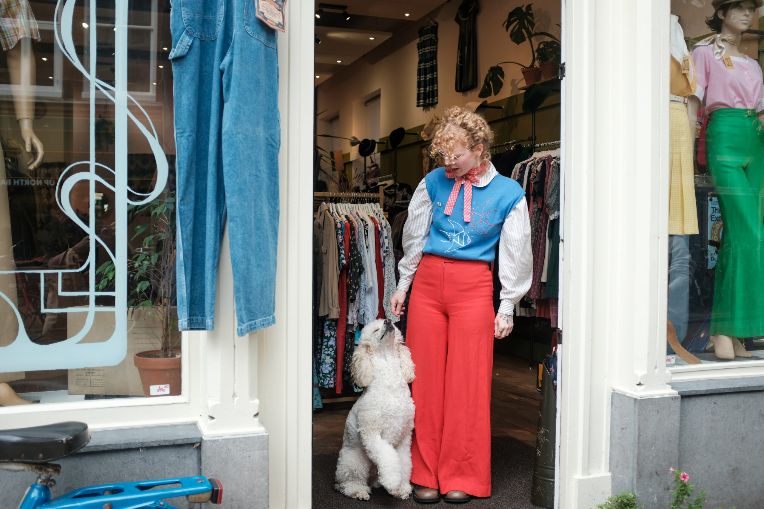 Vrouw met roodblonde krullen en rode broek staat met witte poedel voor een vintagewinkel