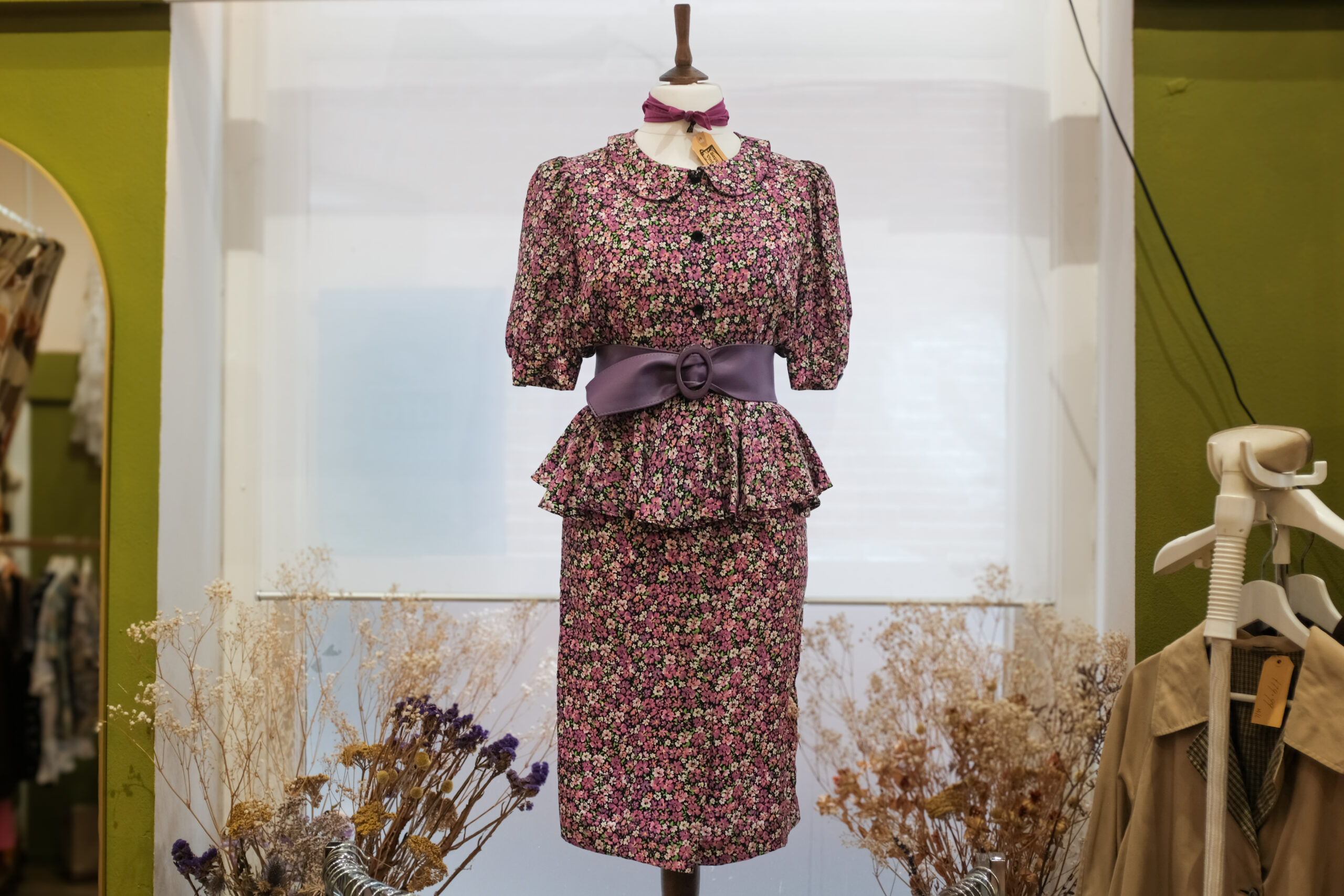 Bloemenjurk met paarse riem op paspop, omringd door droogbloemen