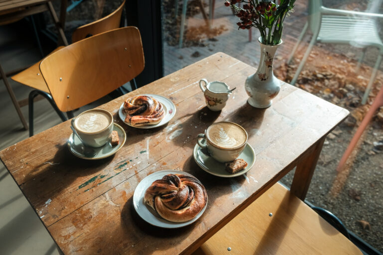 Twee cappuccino’s en kaneelbroodjes op een verweerde houten tafel, met een vaas bloemen en herfstbladeren zichtbaar buiten het raam.