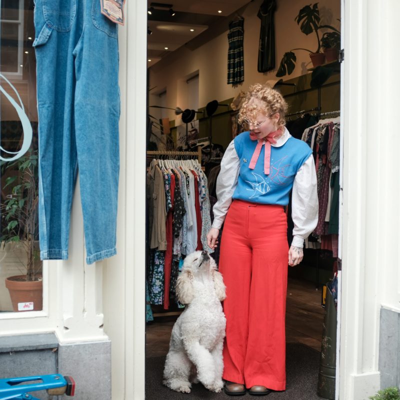 Vrouw met roodblonde krullen en rode broek staat met witte poedel voor een vintagewinkel